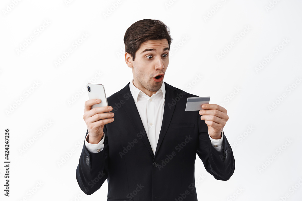 Wow awesome bank. Amazed businessman, corporate man stare amazed at credit card and holding smartphone, standing in black suit against white background Wow awesome bank. Amazed businessman, corporate man stare amazed at credit card and holding smartphone, standing in black suit against white background