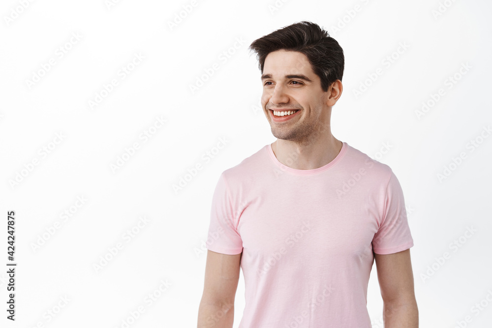 Handsome friendly man in t-shirt looks aside with pleased smile, satisfied with promo, looking at logo and nod in approval, standing over white background Handsome friendly man in t-shirt looks aside with pleased smile, satisfied with promo, looking at logo and nod in approval, standing over white background