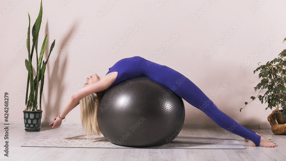 Attractive woman in blue sportswear practices yoga performing bridge pose bandhasana on fitball in the room near the wall foto de Stock Adobe Stock