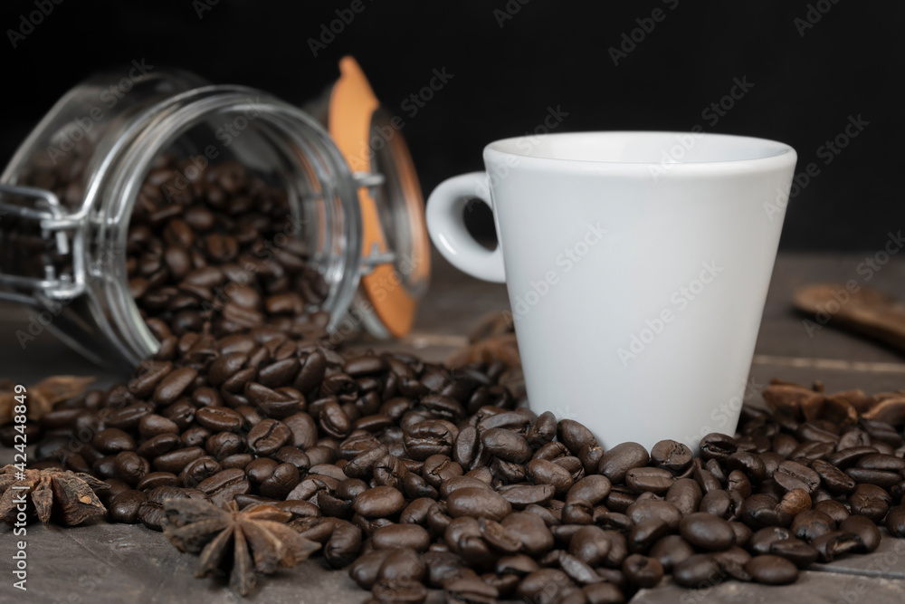 Coffee beans in glass jar with a white coffee cup.Concept of energy caffeine drink on dark background. Coffee beans in glass jar with a white coffee cup.Concept of energy caffeine drink on dark background.