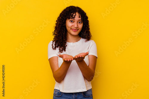 Young mixed race woman isolated on yellow background holding something with palms, offering to camera.