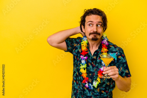 Young caucasian man wearing a hawaiian necklace holding a cocktail isolated on yellow background touching back of head, thinking and making a choice.