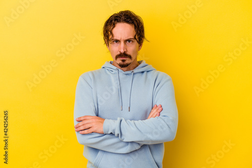 Young caucasian man isolated on yellow background frowning face in displeasure, keeps arms folded. © Asier