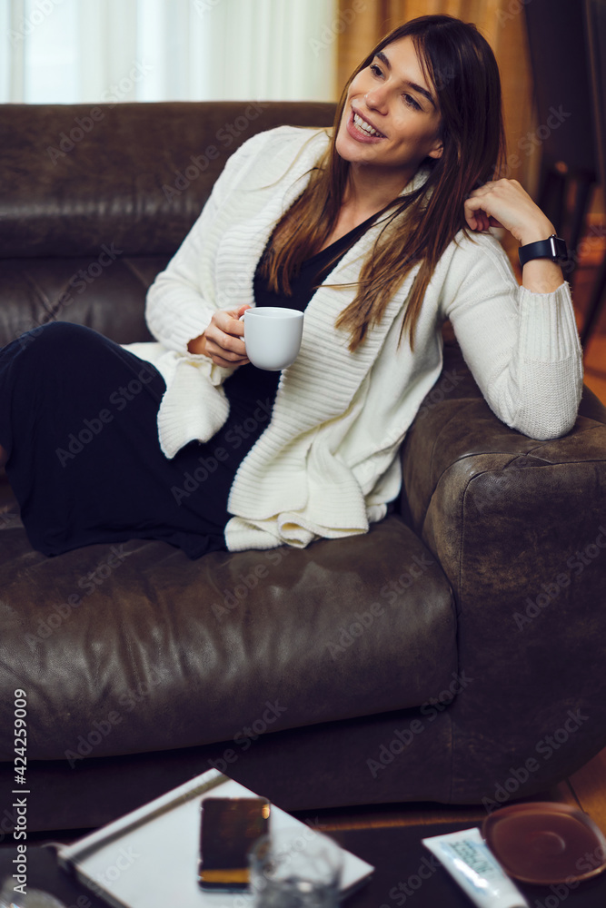 A pregnant woman sitting on a sofa at home with legs crossed and having a coffee break. A pregnant woman sitting on a sofa at home with legs crossed and having a coffee break.