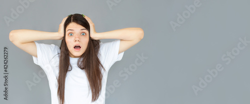 Shocked amazed young woman with hands on head standing and shouting over gray background
