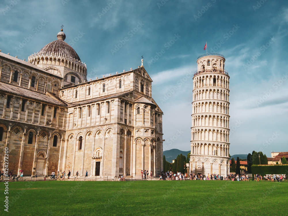 Leaning Tower of Pisa (elaborately adorned 14th-century tower) and Cattedrale di Pisa (marble-striped cathedral) on summer green grass and blue sky. Travel Italy, famous places. Color graded