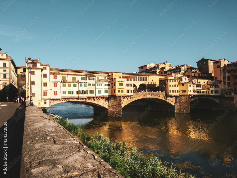 Famous Ponte Vecchio closed-spandrel segmental arch bridge across river Arno at sunset in Florence, Italy. View from embankment with mirror bright reflection in water