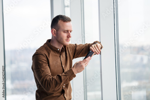 Handsome young male manager stands with the phone near the panoramic window. Male businessman working on a new project