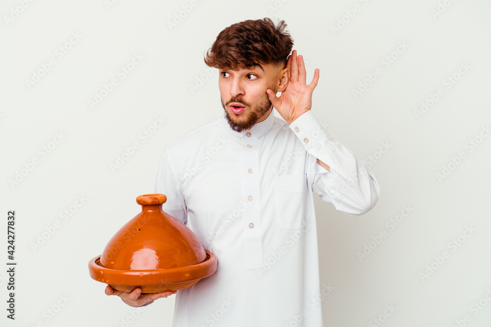 Young Moroccan man wearing the typical arabic costume holding a tajine isolated on white background trying to listening a gossip. Young Moroccan man wearing the typical arabic costume holding a tajine isolated on white background trying to listening a gossip.