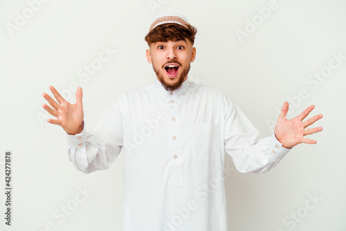 Young Arab man wearing the typical arabic costume isolated on white background receiving a pleasant surprise, excited and raising hands.