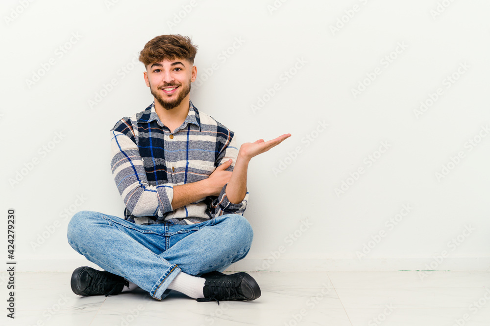Young Moroccan man sitting on the floor isolated on white background showing a copy space on a palm and holding another hand on waist. Young Moroccan man sitting on the floor isolated on white background showing a copy space on a palm and holding another hand on waist.