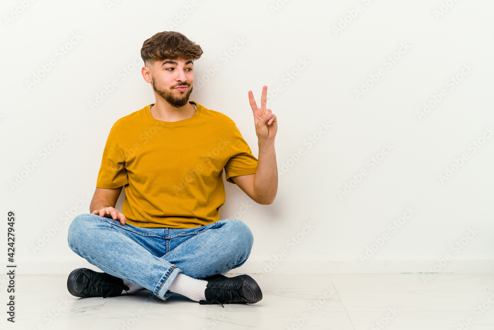 Young Moroccan man sitting on the floor isolated on white background joyful and carefree showing a peace symbol with fingers. Young Moroccan man sitting on the floor isolated on white background joyful and carefree showing a peace symbol with fingers.