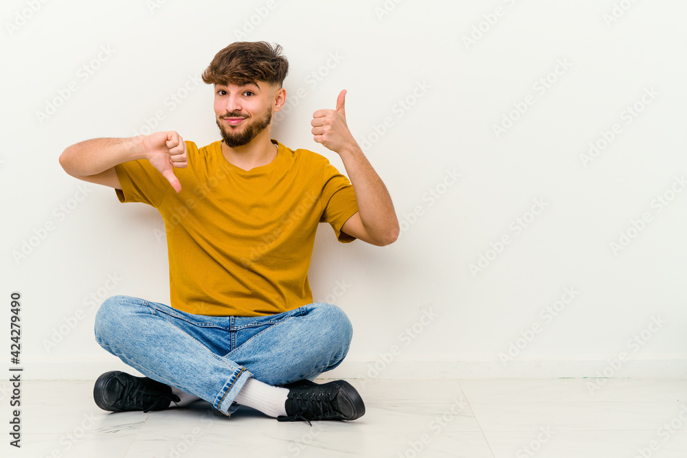 Young Moroccan man sitting on the floor isolated on white background showing thumbs up and thumbs down, difficult choose concept