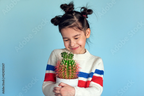 Candid portrait of a pretty smiling positive little girl holds cactus in hands posing on studio blue color background. Cute gardener kid posing with a houseplant.