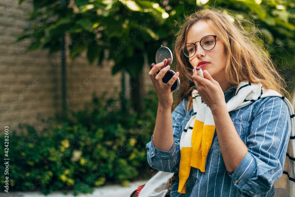 Portrait of a young woman in a casual outfit puting liptisck on her lips, posing outdoors. Pretty female student with long red curly hair walking in the city street on a sunny day.