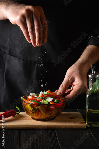 The chef cook sprinkles salted fresh vegetable salad in plate on a wooden table. Cooking healthy food in the kitchen in a restaurant