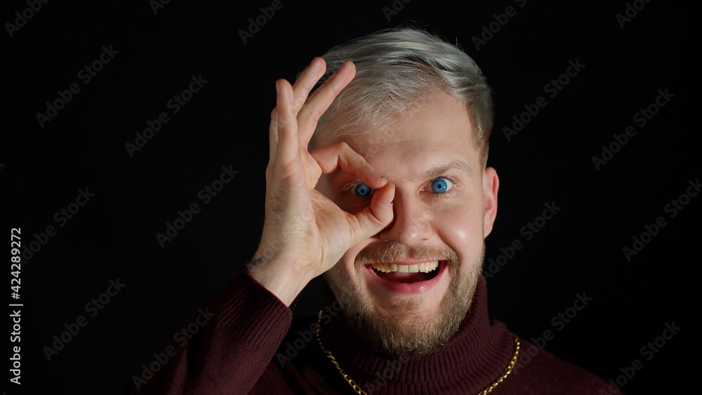 Cheerful excited young man in trendy stylish clothes looking at camera charming smile showing ok okay gesture near his eye against black background. Attractive guy studio shot. Fun and enjoyment