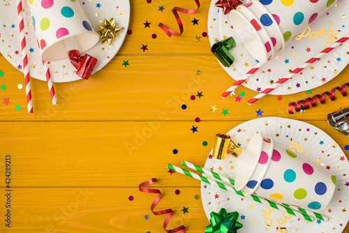 Above photo of cups drink with party tubules whistle multicolored confetti bow and plates isolated on the yellow wooden background with blank space