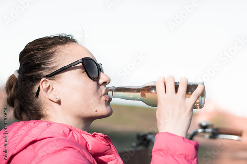 A caucasian woman wearing a pink down jacket and sunglasses drinking beer from a bottle at a picnic. Nature as a background. 