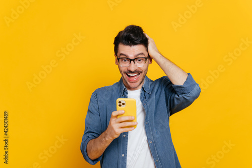 Cheerful excited modern positive caucasian guy uses his smartphone, texting with friends, browsing Internet, social networks, get unexpected news, stands on isolated orange background, smiling