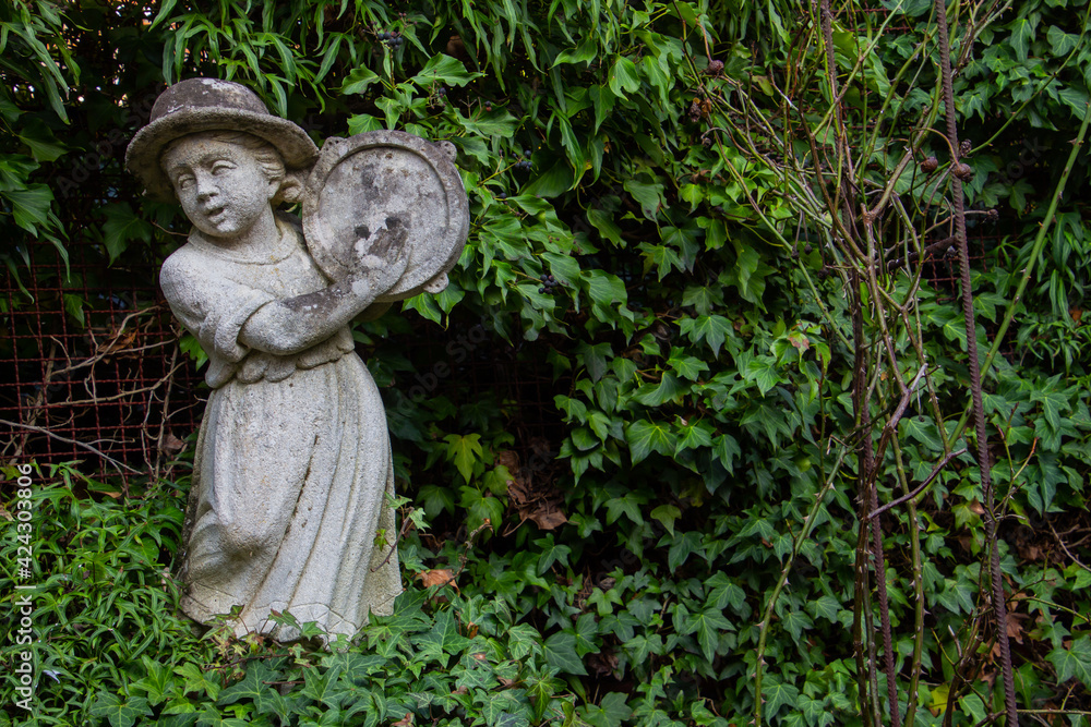 Old white weathered statue of a girl playing a tambourine surrounded with ivy and copy space Old white weathered statue of a girl playing a tambourine surrounded with ivy and copy space