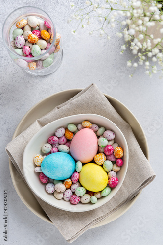 Festive Easter table  with small white flowers, colored eggs and colorful speckled chocolate candies on  ceramic plate. Top view