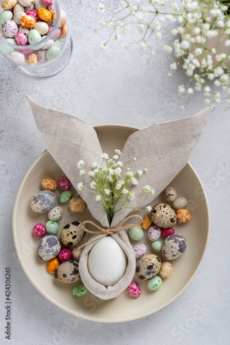 Easter table decoration with egg in napkin in the shape of an Easter bunny with ears, with small white flowers and colorful speckled chocolates on ceramic plate. Top view