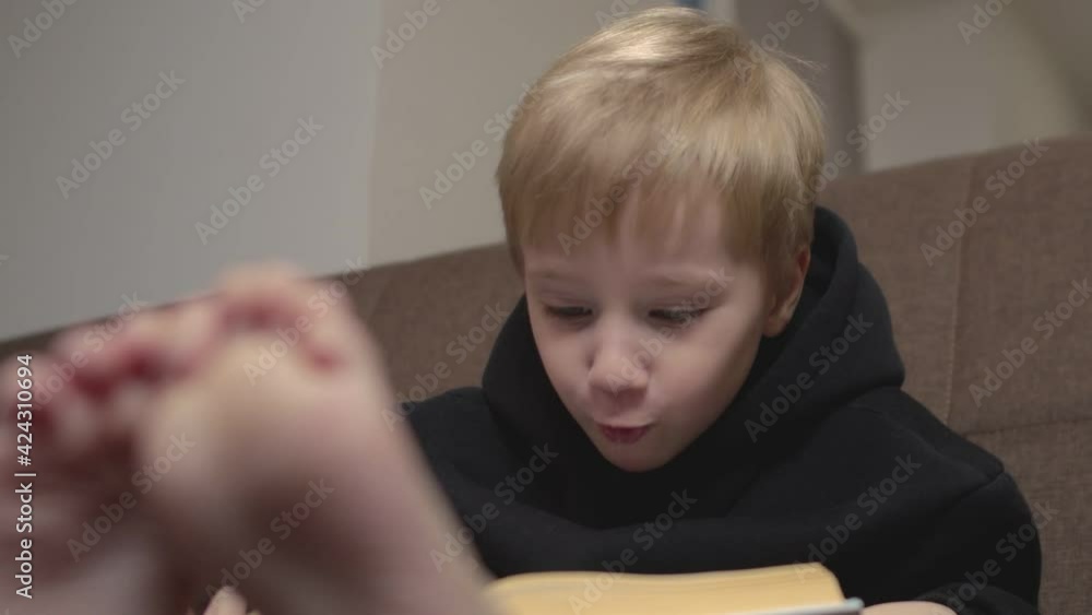Close up of happy little boy in black hoodie sits on couch and reads books. Little boy turns pages on couch Close up of happy little boy in black hoodie sits on couch and reads books. Little boy turns pages on couch