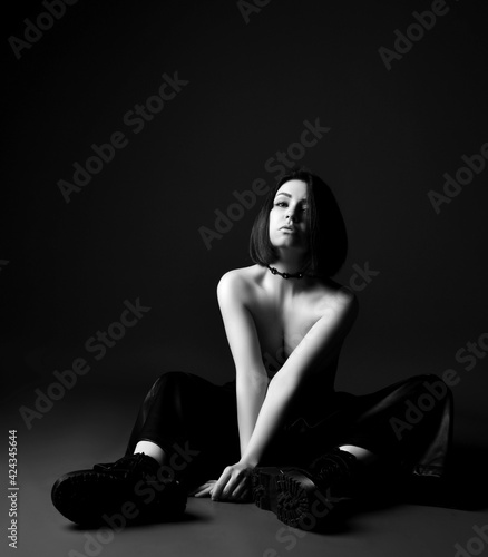 Black and white portrait of young sexy brunette woman in leather pants and brutal choes sitting on floor topless over black background. Fashion, stylish casual look for woman concept photo