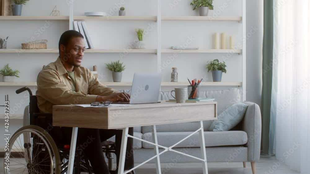Disabled African Guy Working On Laptop Sitting In Wheelchair Indoors Disabled African Guy Working On Laptop Sitting In Wheelchair Indoors