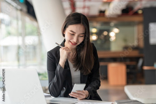 Happy young Asian businesswoman holding a pen using a smartphone at the modern office.