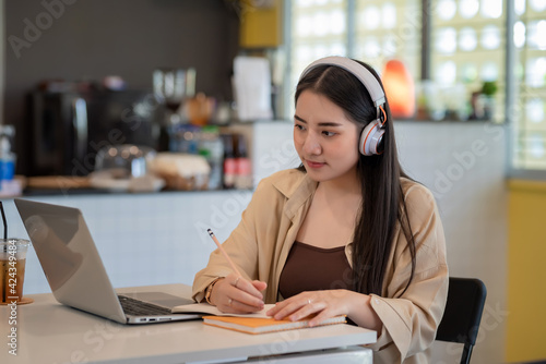 Young Asian woman wearing headphones takes notes and learn online using laptop at café.