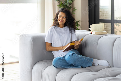Happy woman at home sitting on the couch sitting on the couch reading book. Beautiful African American young woman relaxes reading book at home on sofa