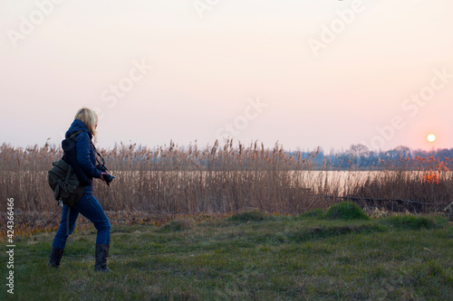 girl photographer on the river bank blonde back view. traveler photographer, blogger, river bank overgrown with grass, setting sun. the concept of tourism, travel. autumn nature