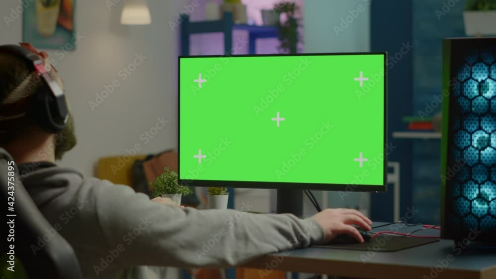 Pro gamer streaming video games with green screen mock-up display in gaming home studio. Player using RGB professional computer with chroma key isolated desktop playing shooter games wearing headset Pro gamer streaming video games with green screen mock-up display in gaming home studio. Player using RGB professional computer with chroma key isolated desktop playing shooter games wearing headset