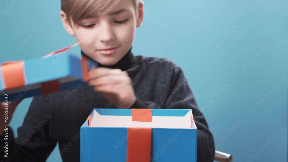 Happy blonde boy with blue eyes opens a gift box on a blue background and is surprised, light is shining from the box. Celebration concept, birthday, new year, christmas. Happy blonde boy with blue eyes opens a gift box on a blue background and is surprised, light is shining from the box. Celebration concept, birthday, new year, christmas.