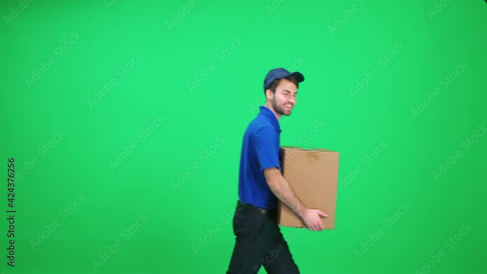 Cheerful man courier in uniform walks with a cardboard box in his hands on a green background, stops and looks at the camera, delivery service, chromakey template. Cheerful man courier in uniform walks with a cardboard box in his hands on a green background, stops and looks at the camera, delivery service, chromakey template.
