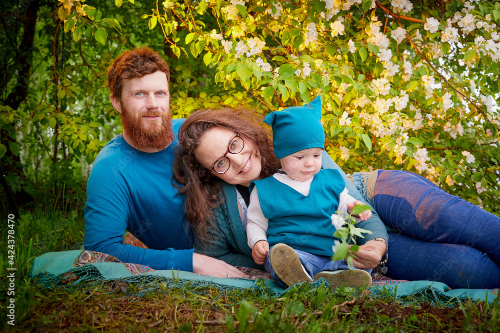 Fototapeta premium Young family with kid having picnic outdoors near apple tree with white flowers. Parents with one child relax in spring garden. Mother, father, little boy in green park.