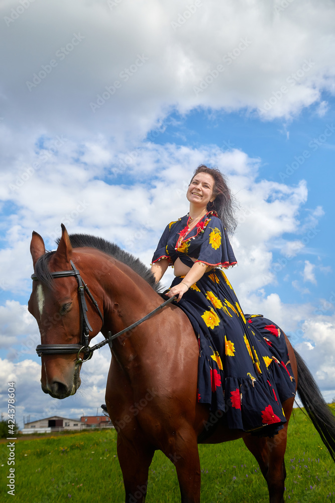 Fototapeta premium Beautiful gypsy girl on a horse in field with green glass in summer day and blue sky and white clouds background. Model in ethnic dress posing with farm animal