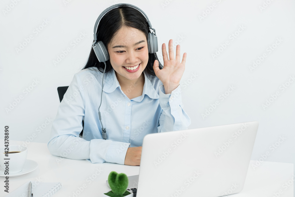 Video call, a young Asian woman wearing headphones, communicates via online video long distance, looking at the digital laptop screen, happily greeted with a friend Video call, a young Asian woman wearing headphones, communicates via online video long distance, looking at the digital laptop screen, happily greeted with a friend