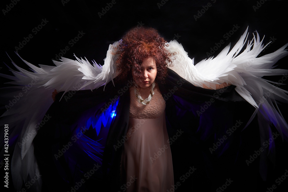 A brunette curly girl in black dress looking like an evil angel with white wings on a black background. Model and actress posing in the studio A brunette curly girl in black dress looking like an evil angel with white wings on a black background. Model and actress posing in the studio