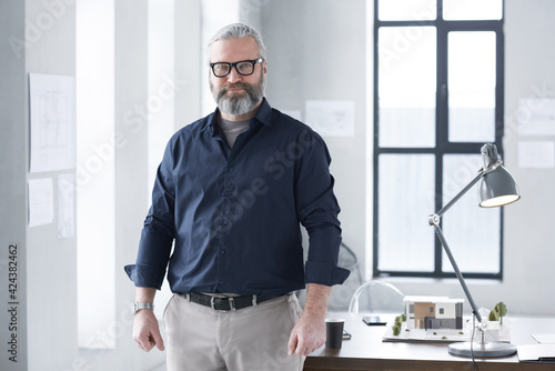 Portrait of mature bearded architect looking at camera while working at office with architectural model