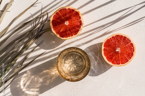 Fototapeta Still life scene with palm leaf, red grapefruit and glass on beige background in sunlight