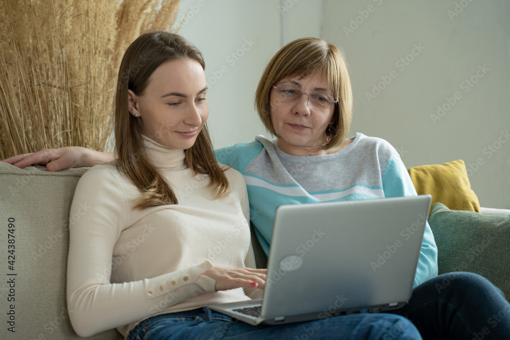 Happy elderly middle mother sitting on couch with her daughter, looking at laptop. Happy mature woman with her adult daughter smiling while sitting on sofa and using laptop in living room