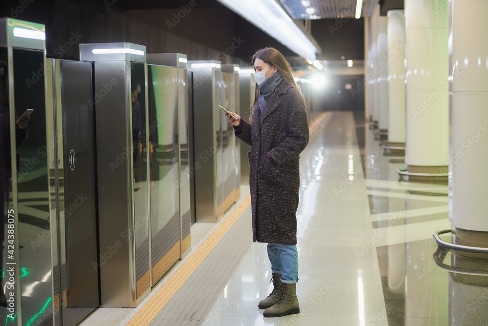 A woman in a medical face mask to avoid the spread of coronavirus is scrolling the news on a smartphone while waiting for a train on the subway. Girl in a surgical mask is keeping social distance.