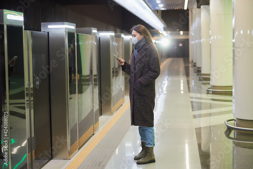 A woman in a medical face mask to avoid the spread of coronavirus is scrolling the news on a smartphone while waiting for a train on the subway. Girl in a surgical mask is keeping social distance.