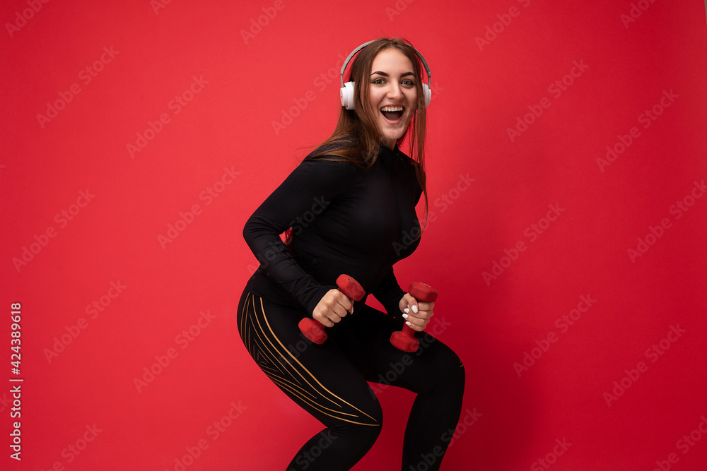 Side-profile photo of beautiful happy smiling young brunet woman wearing black sport clothes isolated on red background wall squating using dumbbells wearing white headsets listening to music looking Side-profile photo of beautiful happy smiling young brunet woman wearing black sport clothes isolated on red background wall squating using dumbbells wearing white headsets listening to music looking