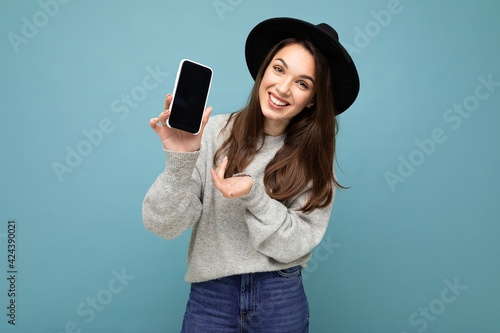 Pretty young smiling woman wearing black hat and grey sweater holding phone looking at camera isolated on background photo