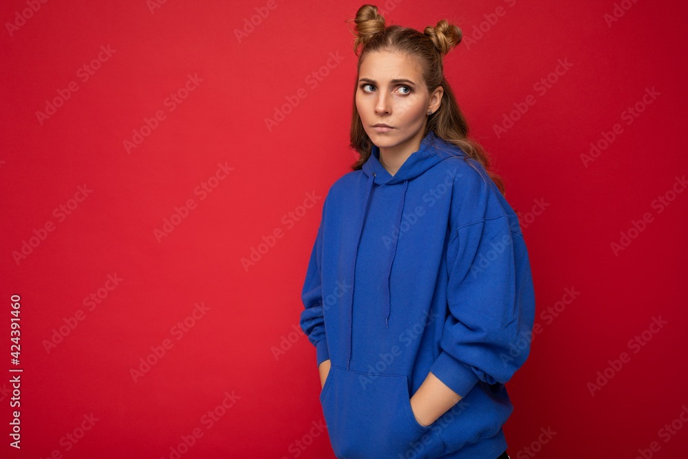 Portrait of young sad upset attractive winsome blonde woman with two horns with sincere emotions wearing casual bright blue hoodie isolated on red background with free space