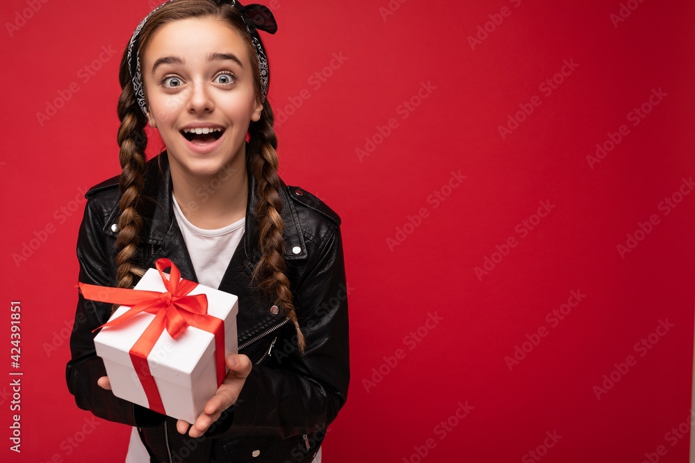 Photo of beautiful happy surprised brunette girl isolated over red background wall wearing white casual t-shirt and stylish black leather jacket holding white gift box with red ribbon and looking at Photo of beautiful happy surprised brunette girl isolated over red background wall wearing white casual t-shirt and stylish black leather jacket holding white gift box with red ribbon and looking at